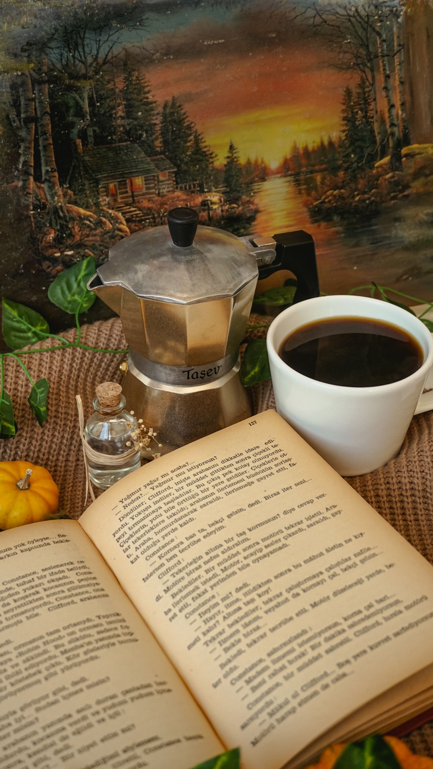 An open book on a table beside a moka pot and a cup of coffee
